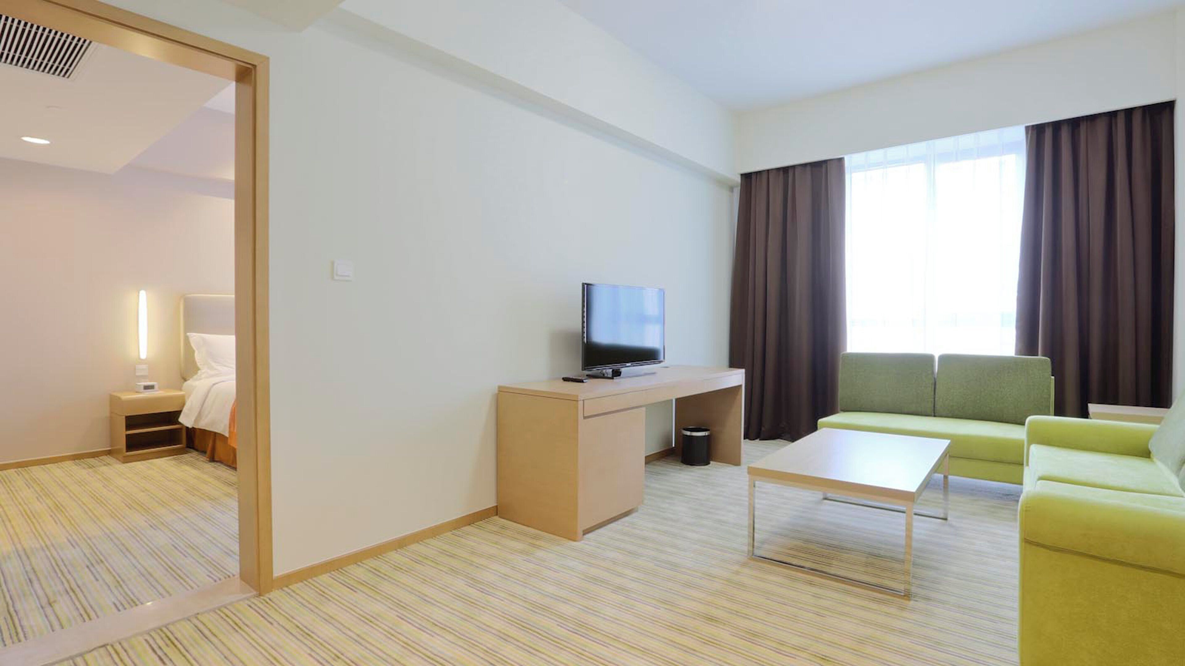 A hotel room with a bed, a desk with a TV, a sofa, and a window with curtains.