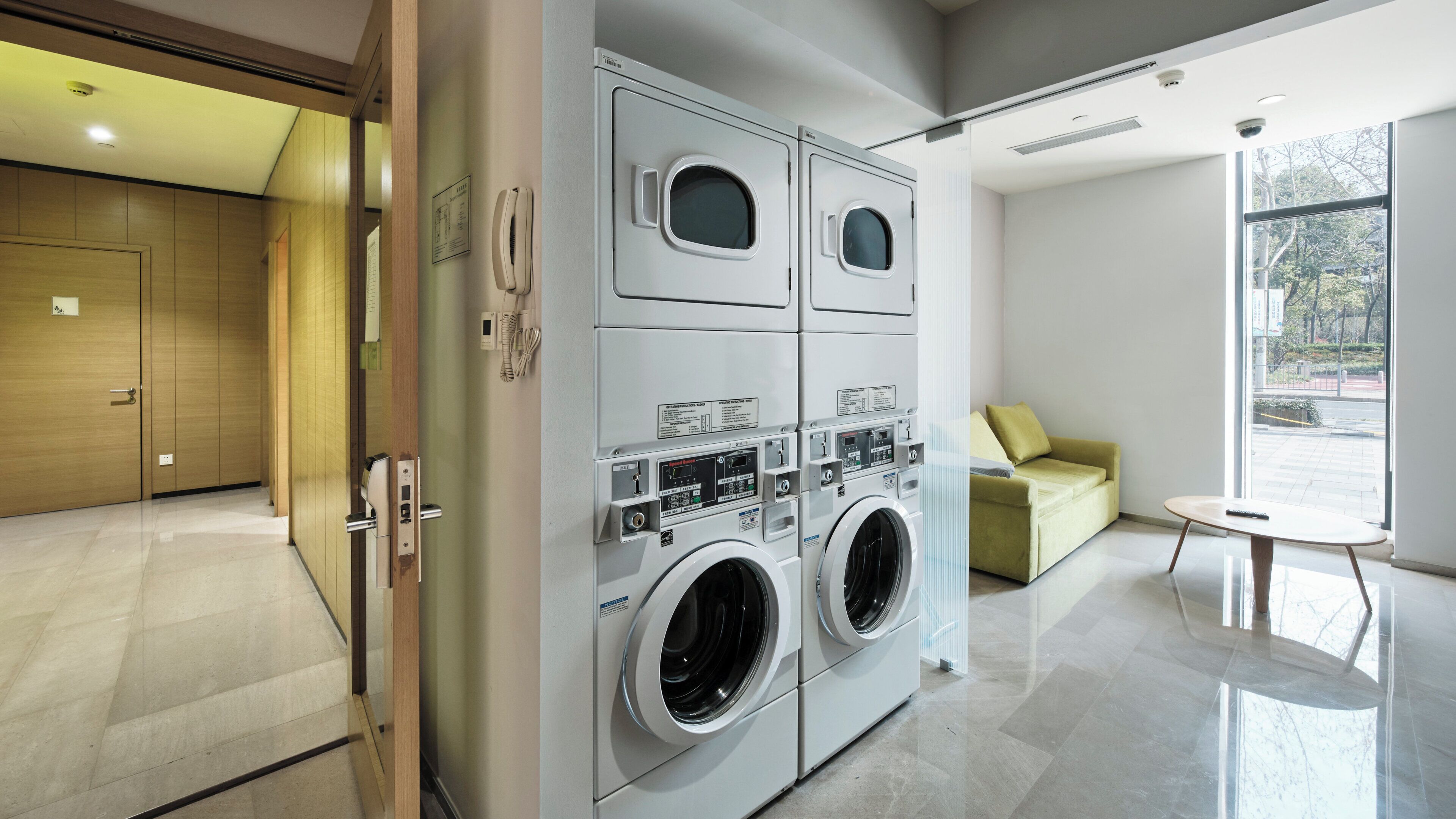 A modern laundry room with two large front-loading washers and dryers, a wooden door, a small table, and a sofa with a view of the outdoors.