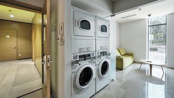 A modern laundry room with two large front-loading washers and dryers, a wooden door, a small table, and a sofa with a view of the outdoors.