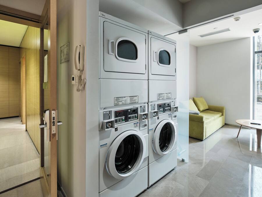A modern laundry room with two large front-loading washers and dryers, a wooden door, a small table, and a sofa with a view of the outdoors.