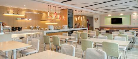 A modern restaurant with multiple dining tables and chairs, a bar area, and a television screen mounted on the wall.