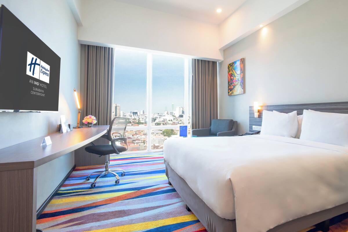 A hotel room with a bed, a desk, a chair, a large window with a city view, and a painting on the wall.