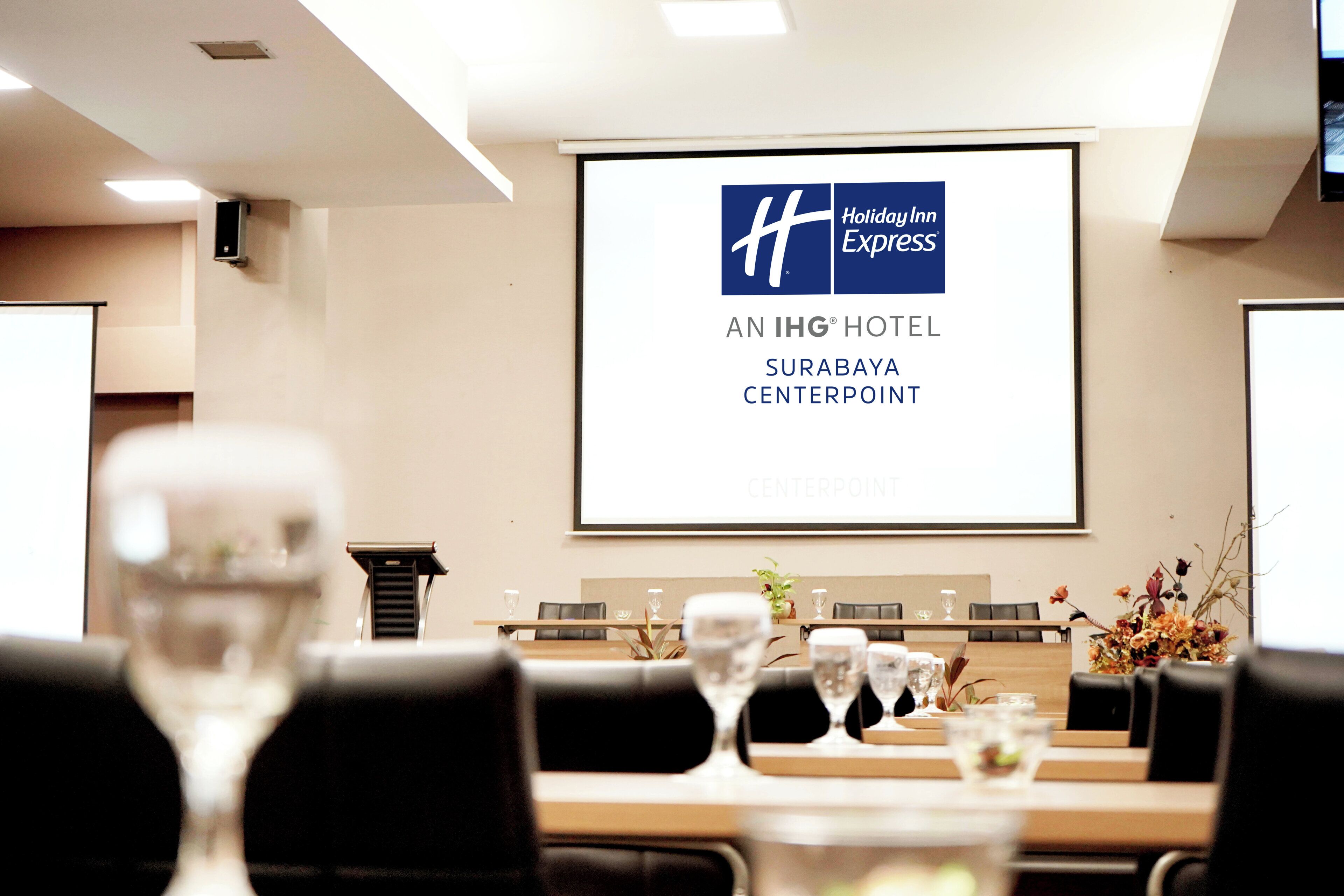 Un salón de conferencias con una pantalla grande mostrando el logotipo de Holiday Inn Express y el texto 'AN IHG HOTEL SURABAYA CENTERPOINT'.