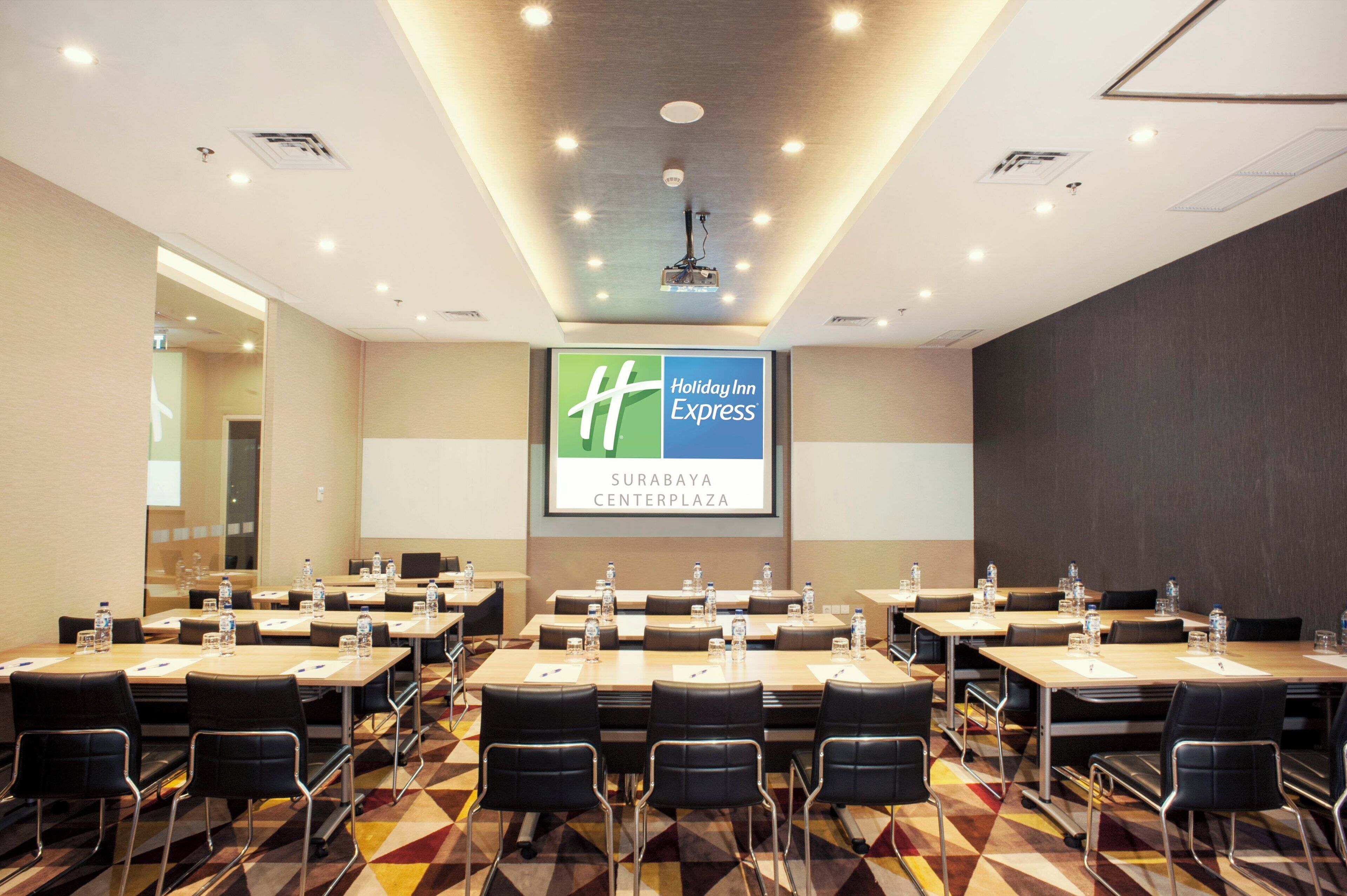 A conference room with multiple tables and chairs, a large screen displaying the Holiday Inn Express logo, and a patterned carpet.