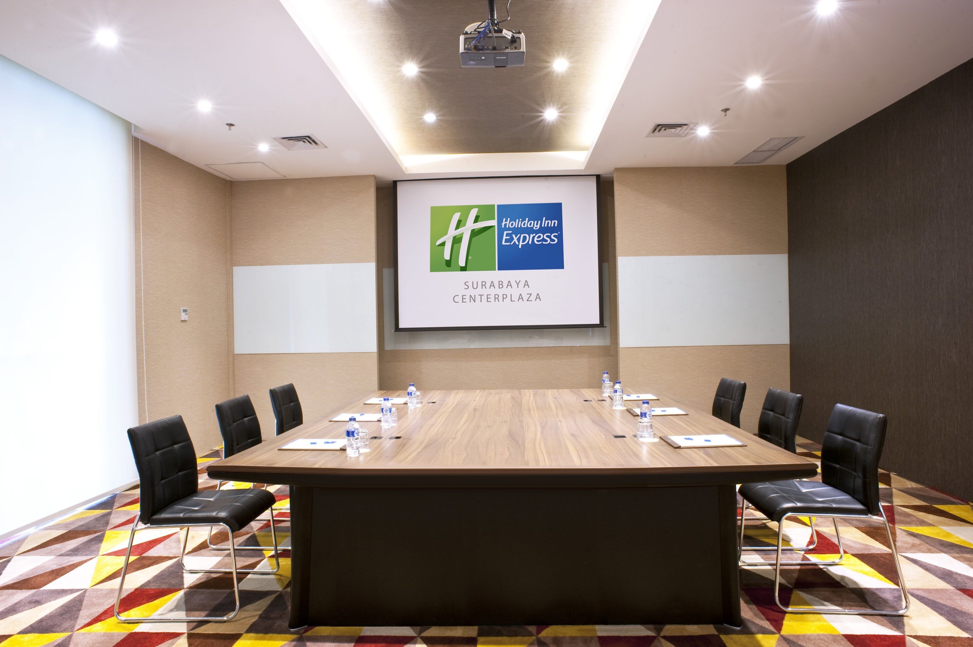 A conference room with a long table, black chairs, a projector screen, and a Holiday Inn Express logo on the wall.