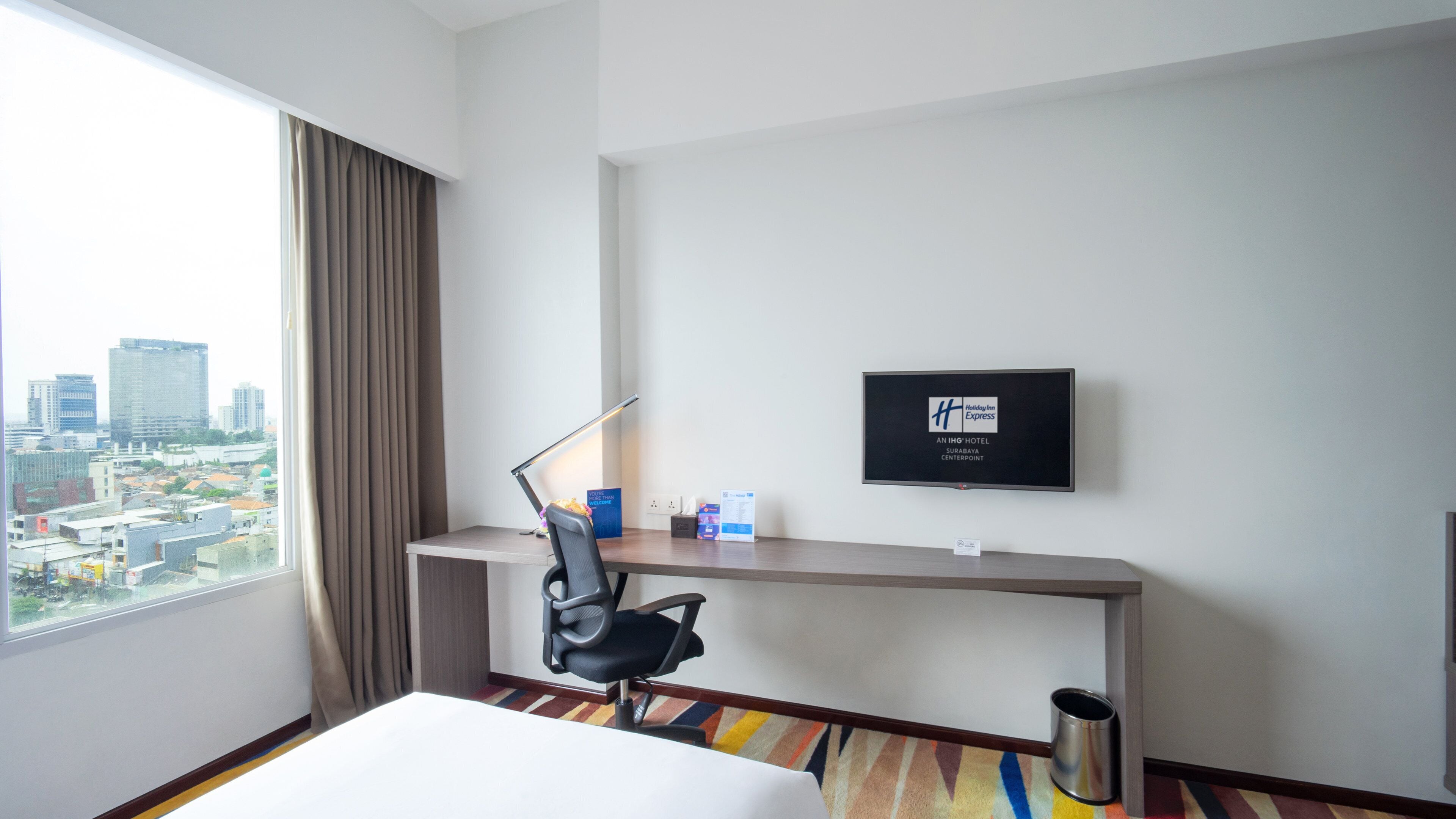 A hotel room with a desk, chair, and a large window offering a city view.