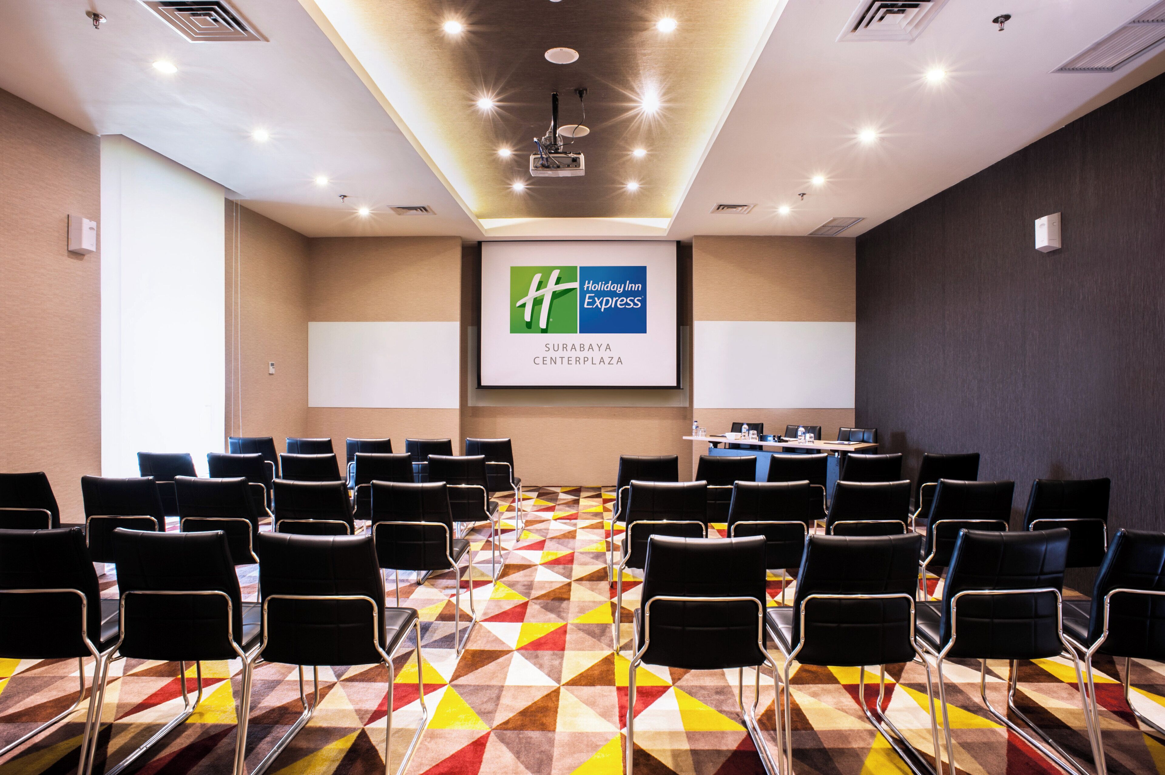 Een vergaderruimte met een groot scherm waarop het logo van Holiday Inn Express staat, geordende stoelen en een kamerbreed tapijt met een patroon.