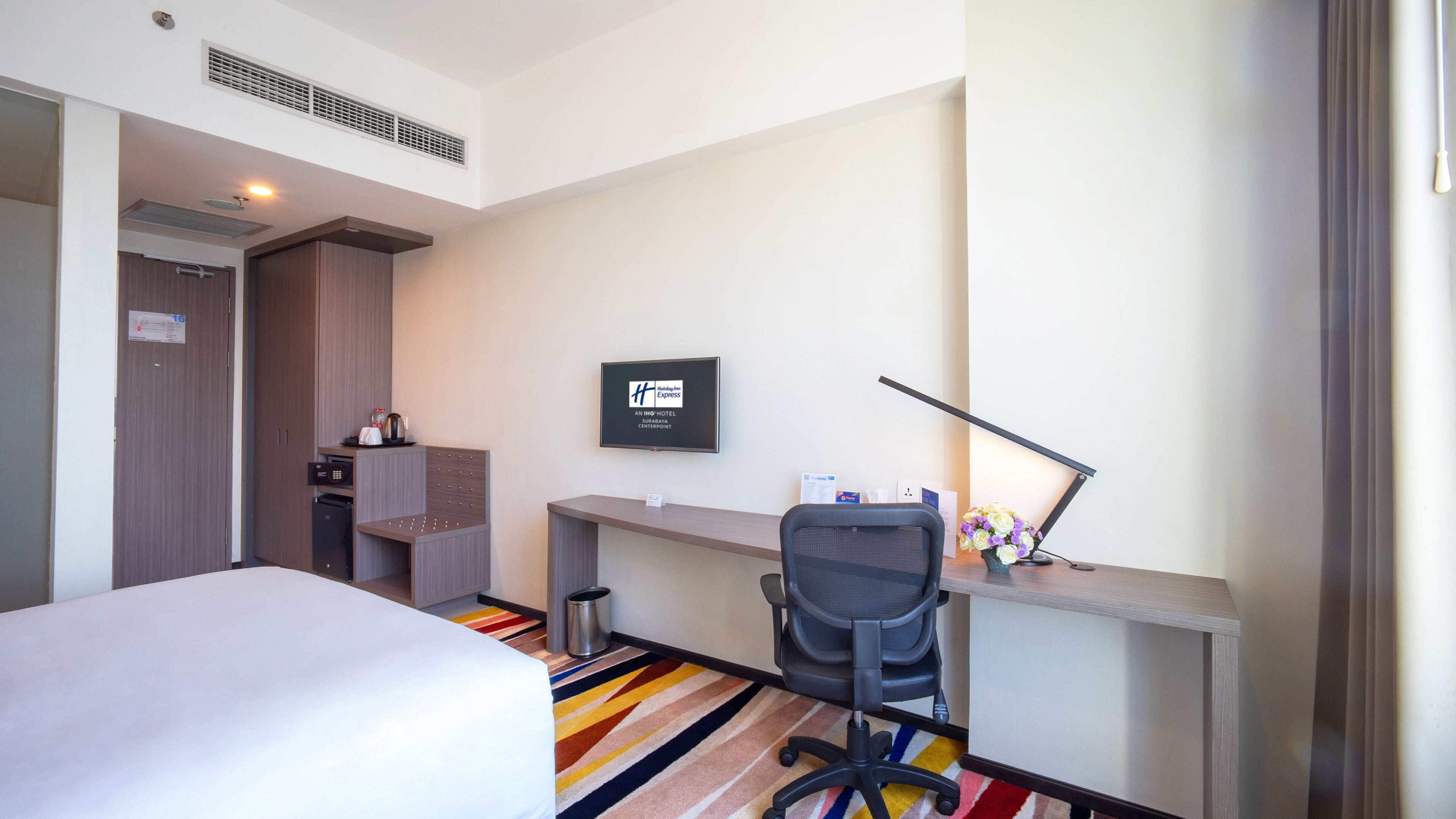 A hotel room with a bed, desk, chair, and a wall-mounted TV.