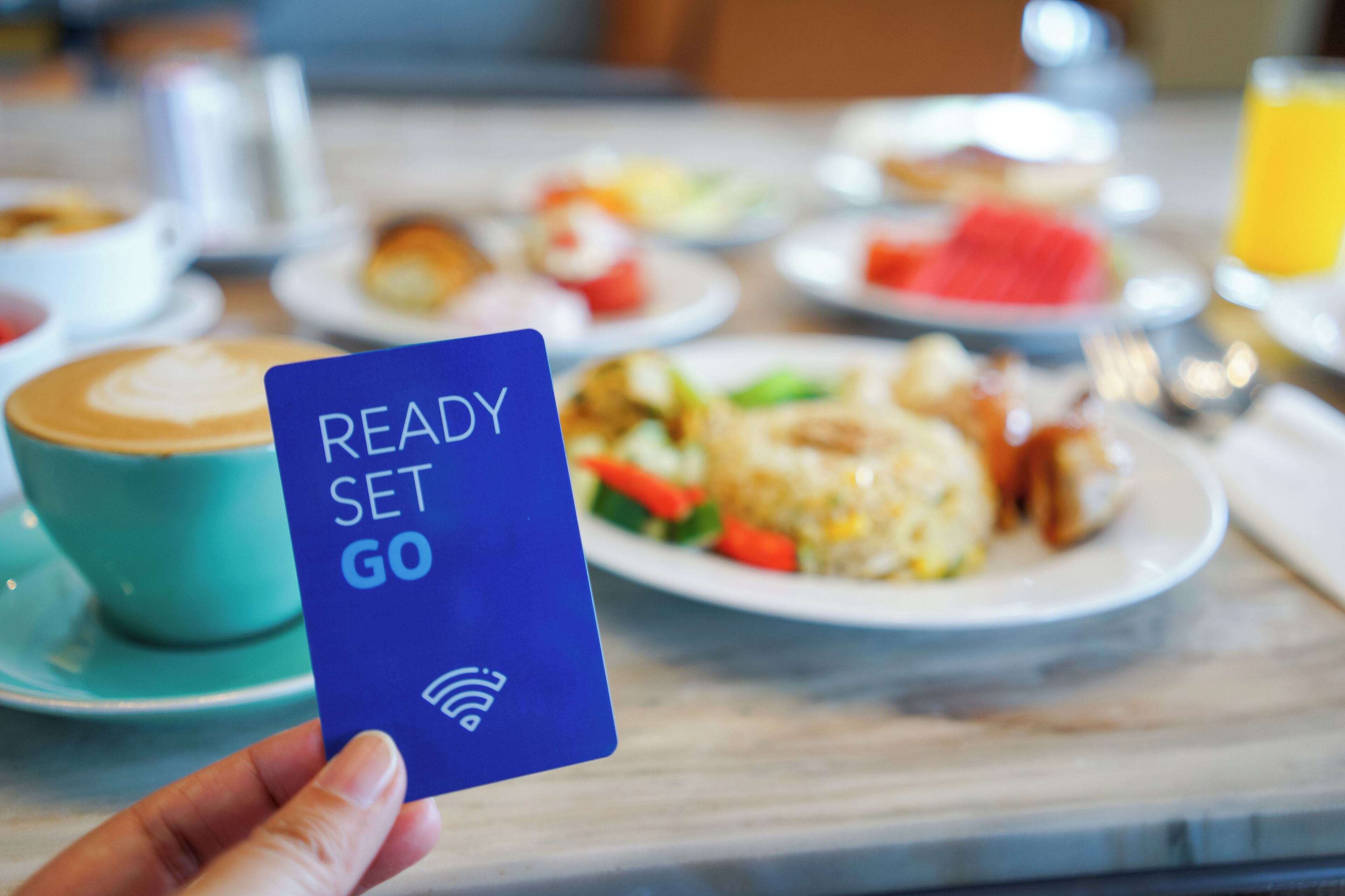 A hand holding a blue card with 'READY SET GO' text, a cup of coffee, and plates of food on a table.