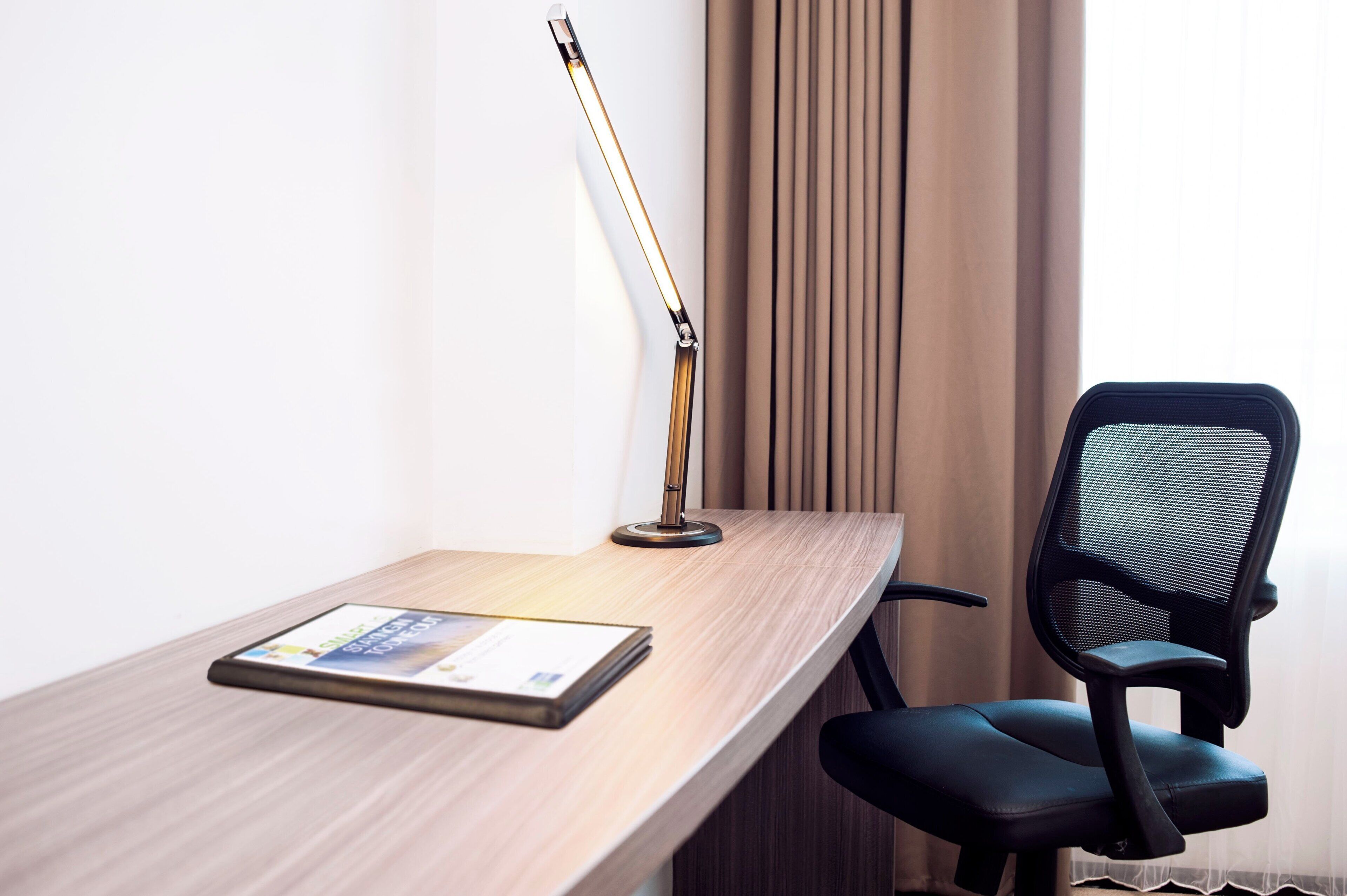 A hotel workspace with a wooden desk, a black office chair, a desk lamp, and a brochure.
