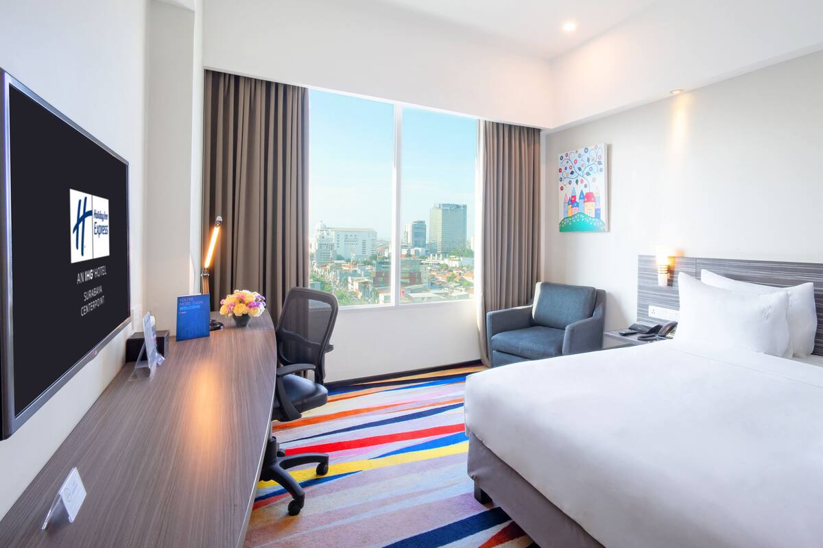 A hotel room with a bed, a desk with a computer, a chair, a nightstand with a phone, and a large window with a city view.