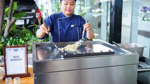 En person laver mad på en stor rustfri griddle, med et skilt der siger "NASI GORENG" og et træbord med tilbehør og en skål i forgrunden.