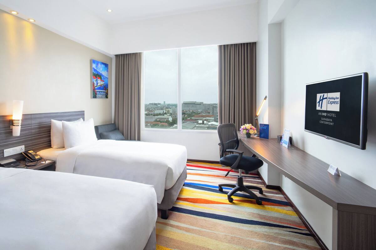 A hotel room with two beds, a desk, a chair, a TV, and a large window with a city view.