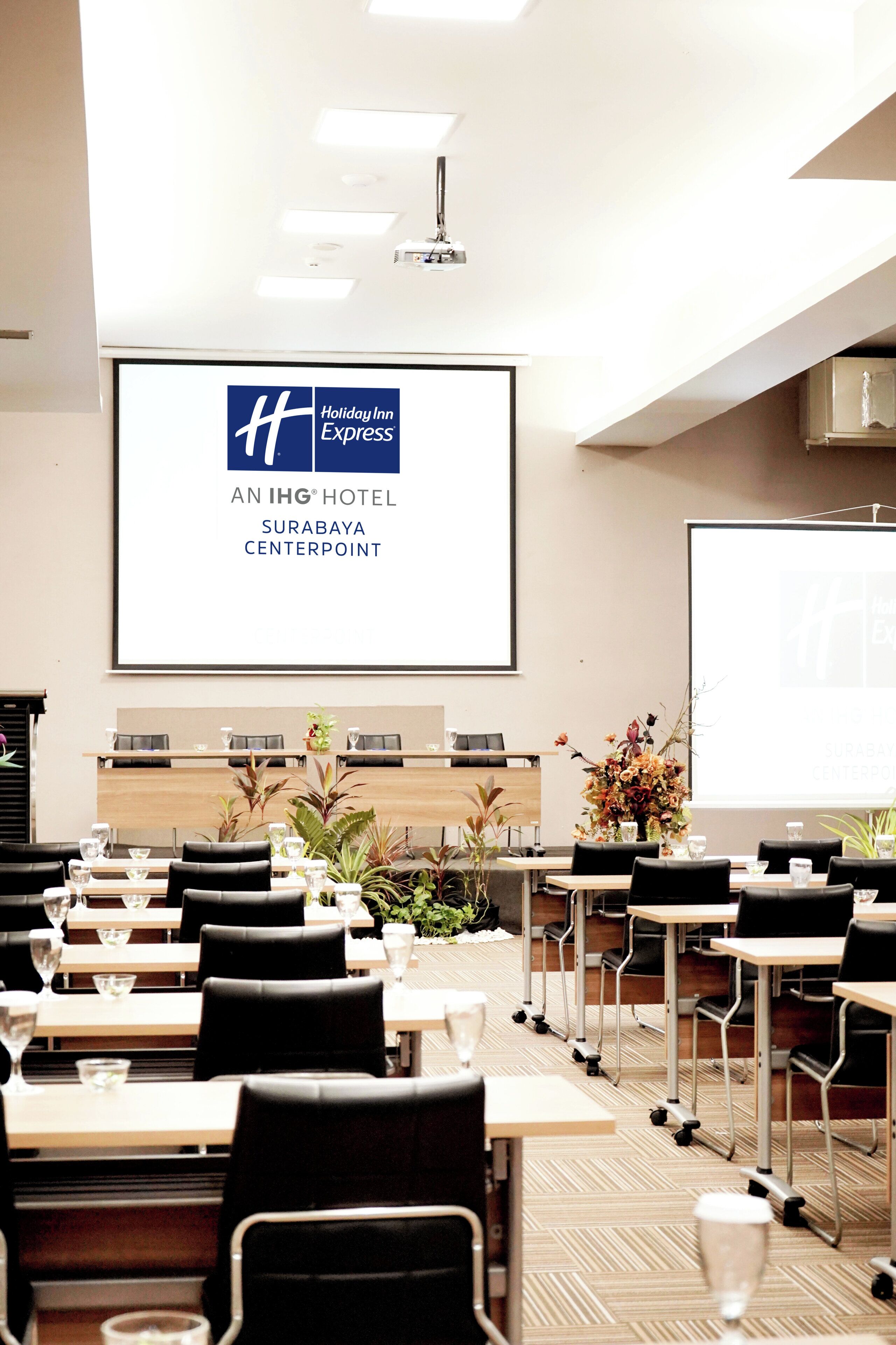 Un salón de conferencias con mesas y sillas, una pantalla de proyector y el logotipo de Holiday Inn Express.