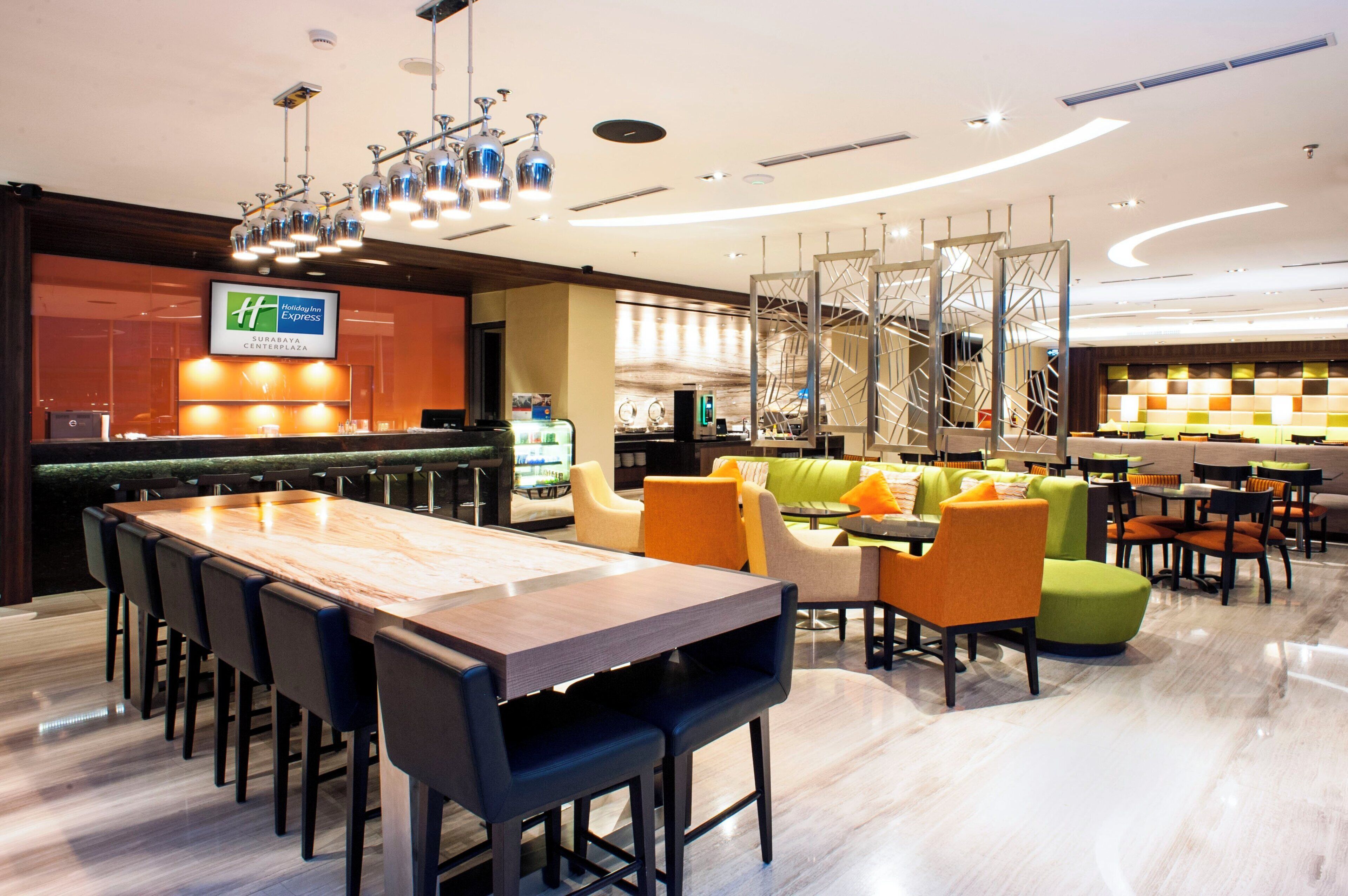 A hotel lobby with a reception desk, a long communal table, and several seating areas with chairs and sofas.