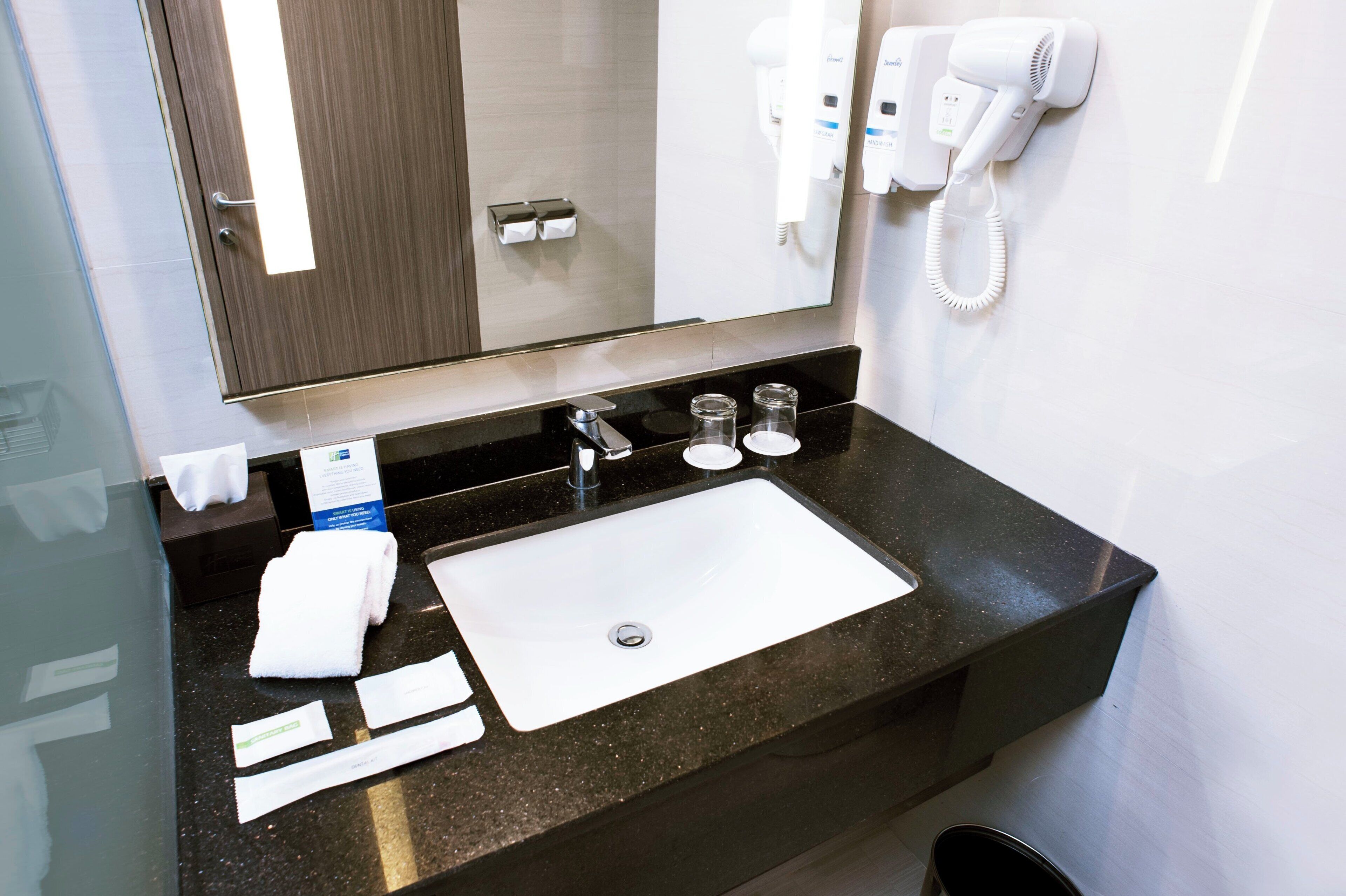 A hotel bathroom with a black countertop, a white sink, a mirror, a hairdryer, and a hand dryer.
