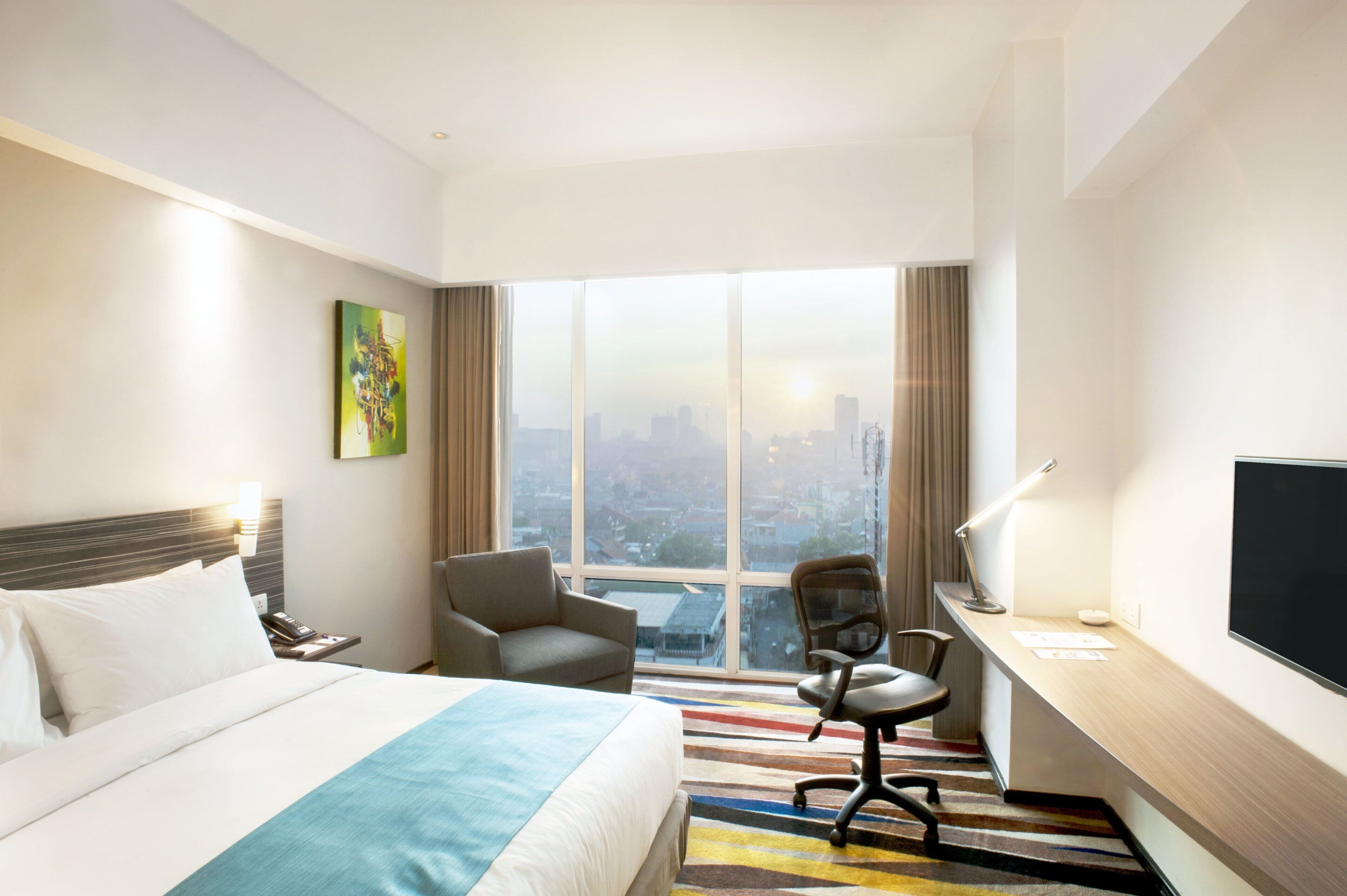 A hotel room with a large bed, a desk with a chair, a TV, and a city view.