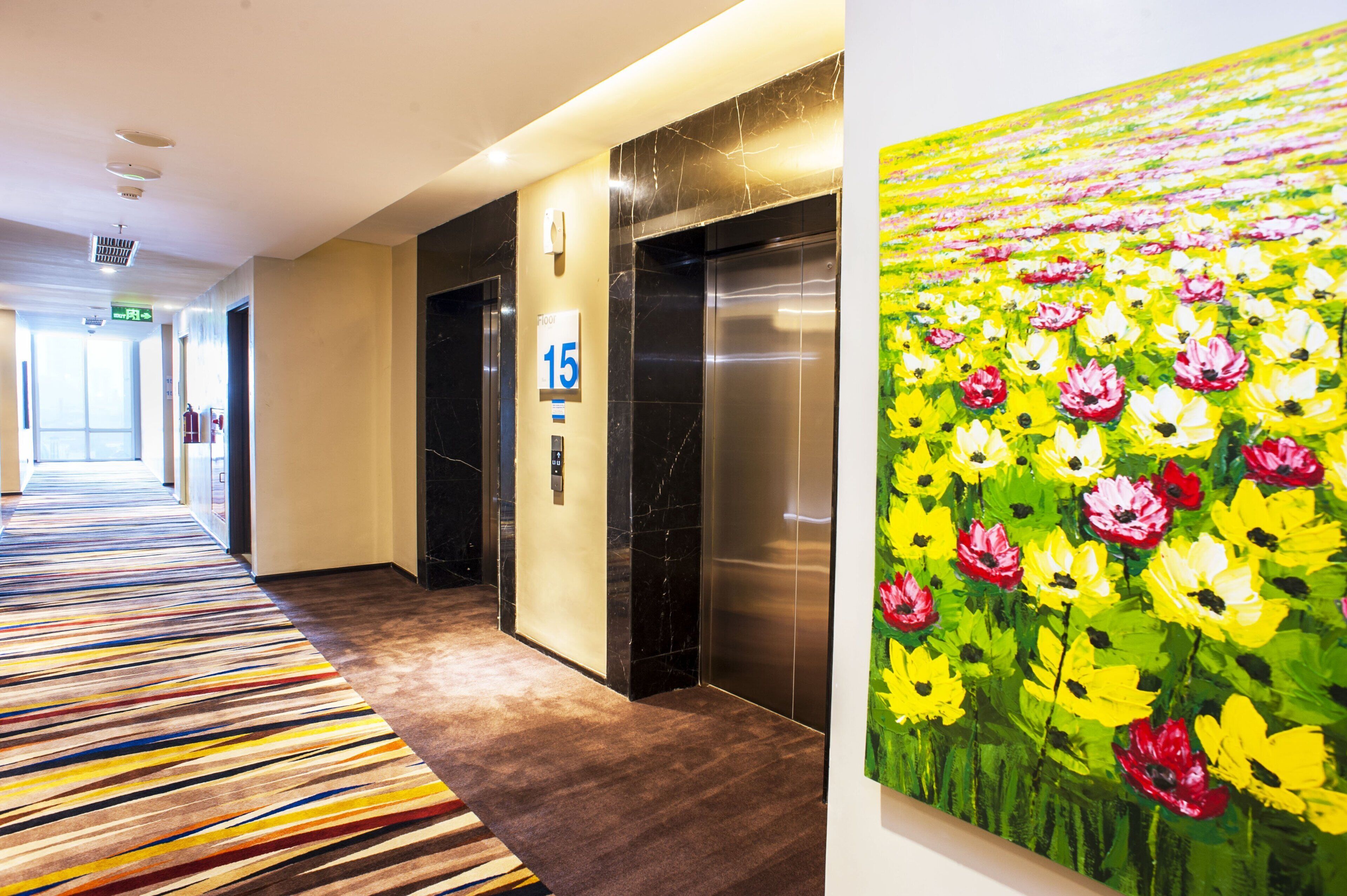 A hotel corridor with a striped carpet, elevators, and a large floral painting on the wall.