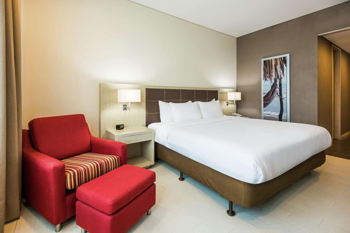 A hotel room with a large bed, a red armchair, a small table with a lamp, and a wall-mounted artwork.