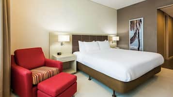 Une chambre d’hôtel avec un grand lit, un fauteuil rouge, une petite table avec une lampe et une œuvre d’art accrochée au mur.