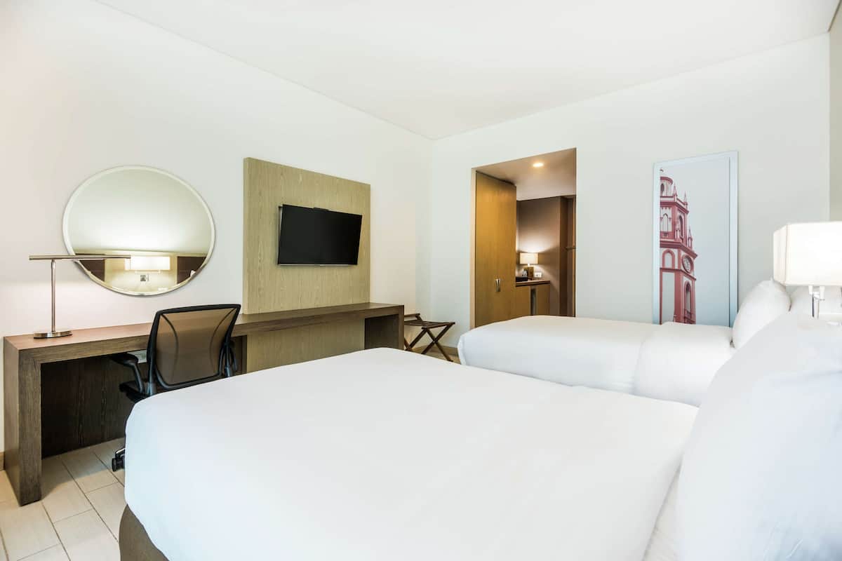 A hotel room with two beds, a desk with a chair, a TV mounted on the wall, and a door leading to another room.