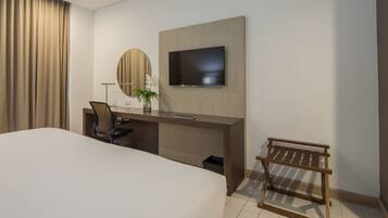 A hotel room with a large bed, a desk with a chair, a television mounted on the wall, a small round mirror, and a potted plant.