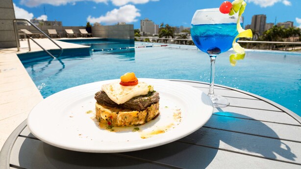 A dining table with a plate of food and a cocktail glass by a poolside. - Hilton Garden Inn Barranquilla (Barranquilla)
