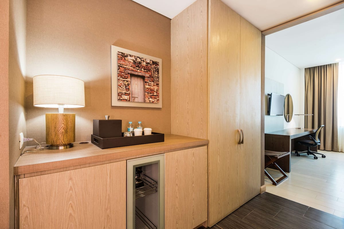 A hotel room with a wooden vanity, a lamp, a small refrigerator, a desk with a chair, a television, and a wall-mounted artwork.