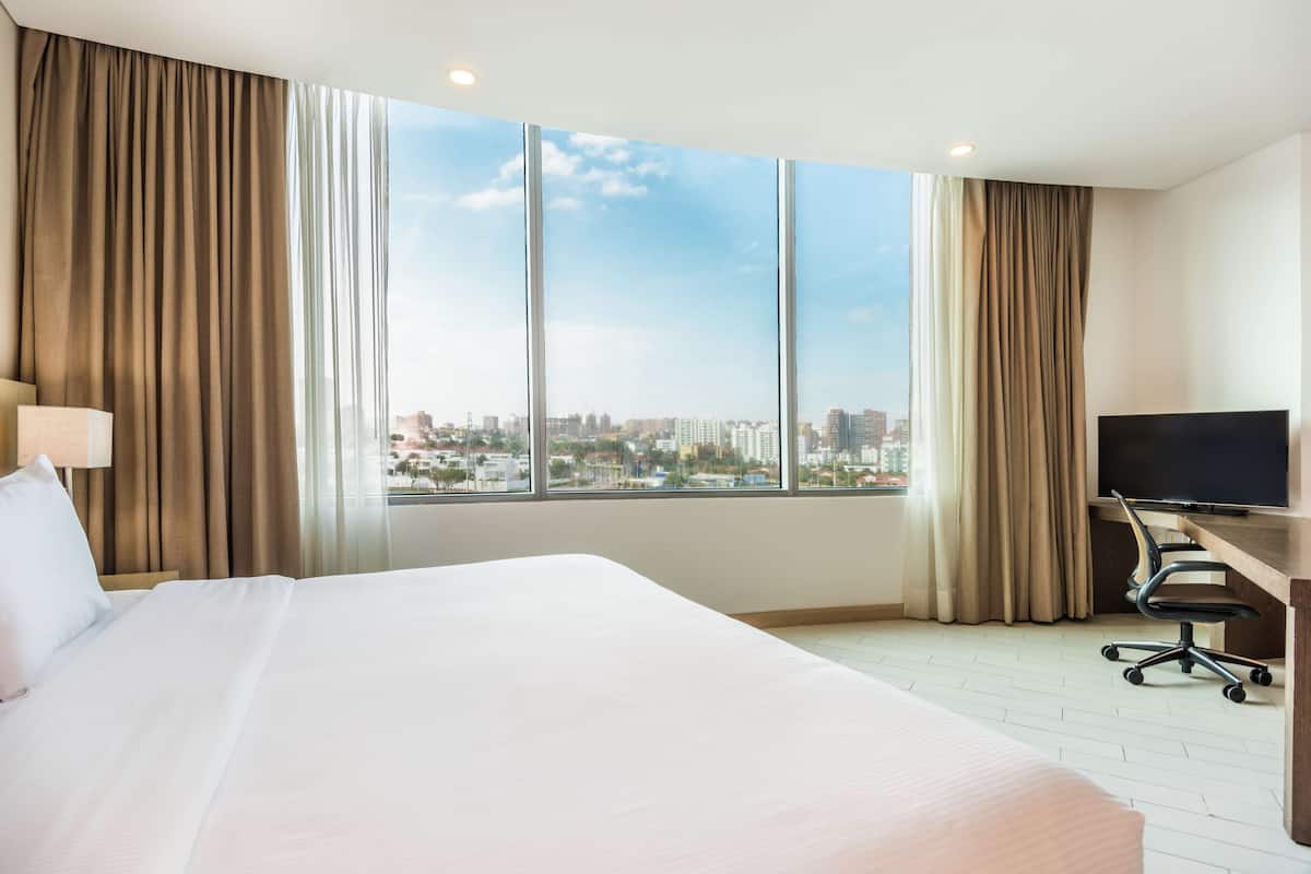 A hotel room with a large bed, a desk with a chair, a television, and a city view through large windows.