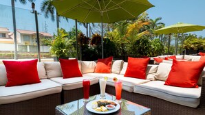 A sunlit outdoor seating area with wicker furniture, red cushions, and a glass table with a plate of food and drinks. - Hilton Garden Inn Barranquilla (Barranquilla)