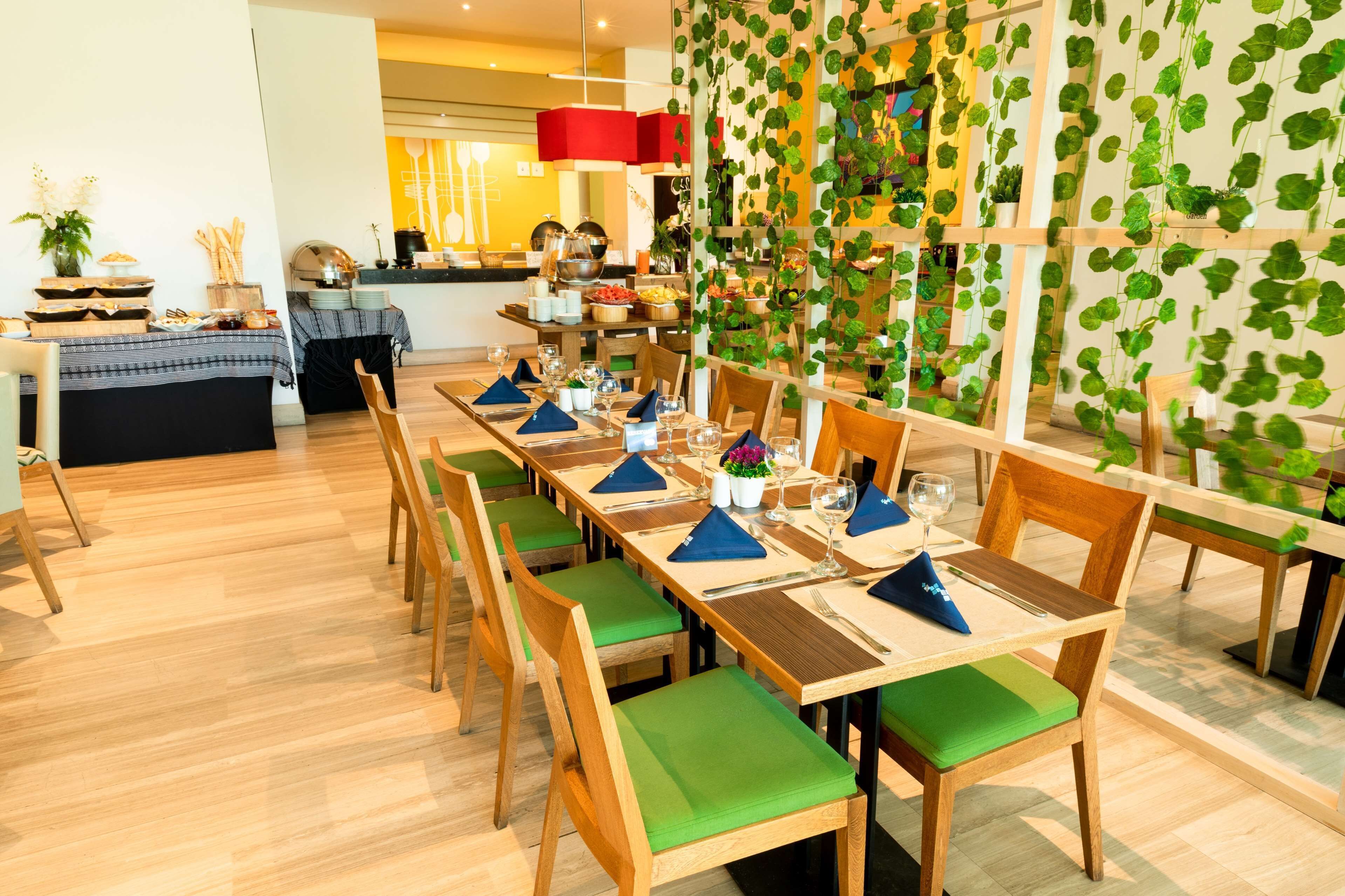 Un comedor con mesas de madera y sillas con cojines verdes, una cocina al fondo y una pared decorada con plantas colgantes.