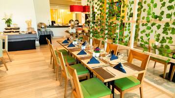 Un comedor con mesas de madera y sillas con cojines verdes, una cocina al fondo y una pared decorada con plantas colgantes.