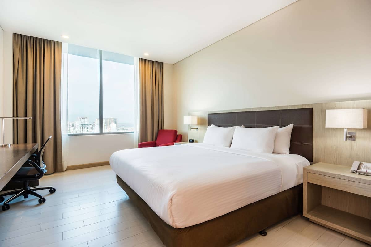 A hotel room with a large bed, a desk with a chair, a lamp, and a view of the city through the window.
