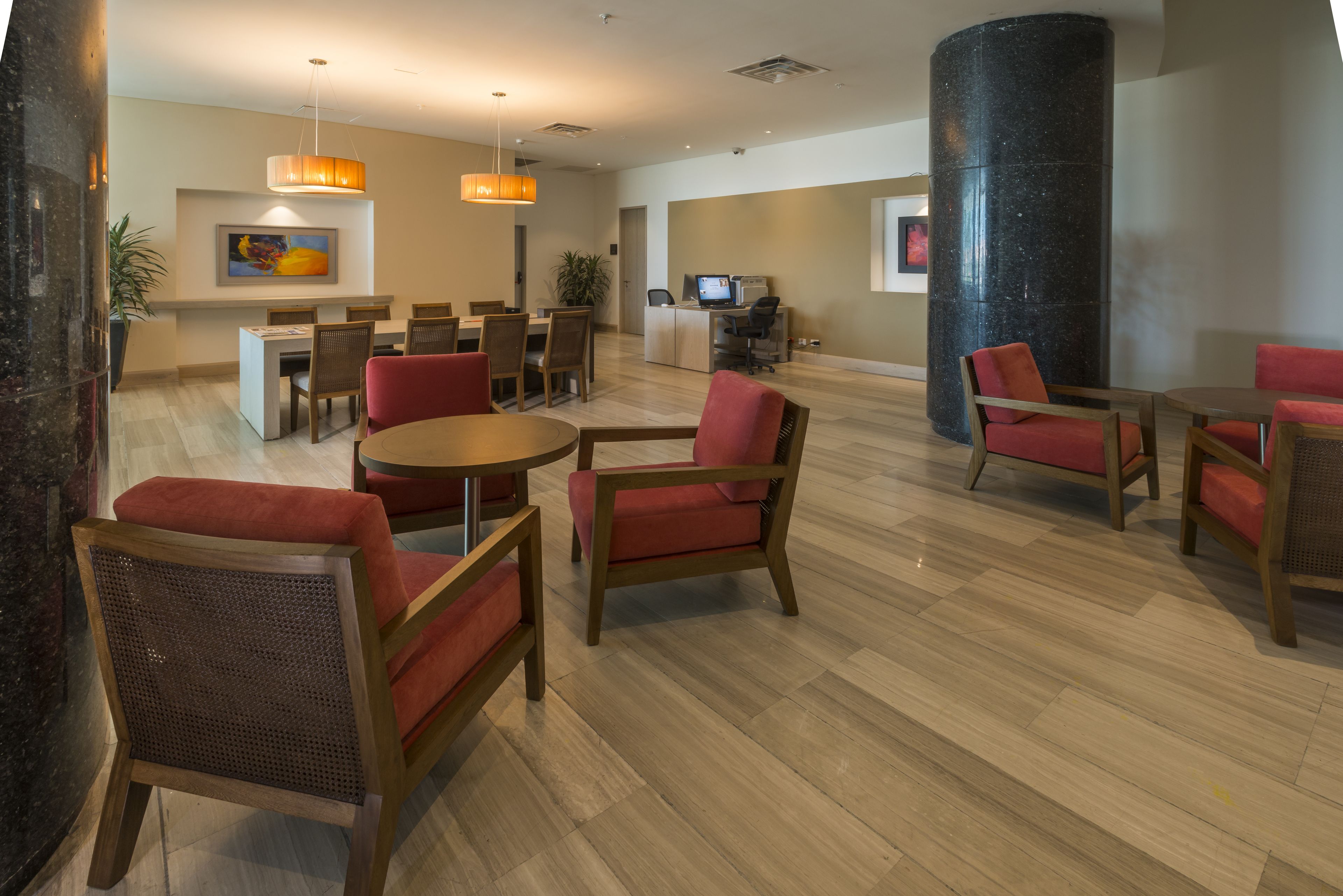 A spacious lobby with a reception desk, multiple seating areas with red chairs, a large black pillar, and a television screen.