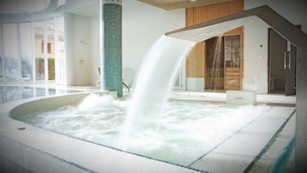 A hot tub with a waterfall feature, surrounded by a tiled floor and a wooden bench. - Thalassa Sport (Roses)