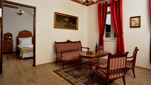 Una habitación con una cama, un banco de madera, una mesa con sillas, un tapete, un cuadro y una ventana con cortinas rojas.