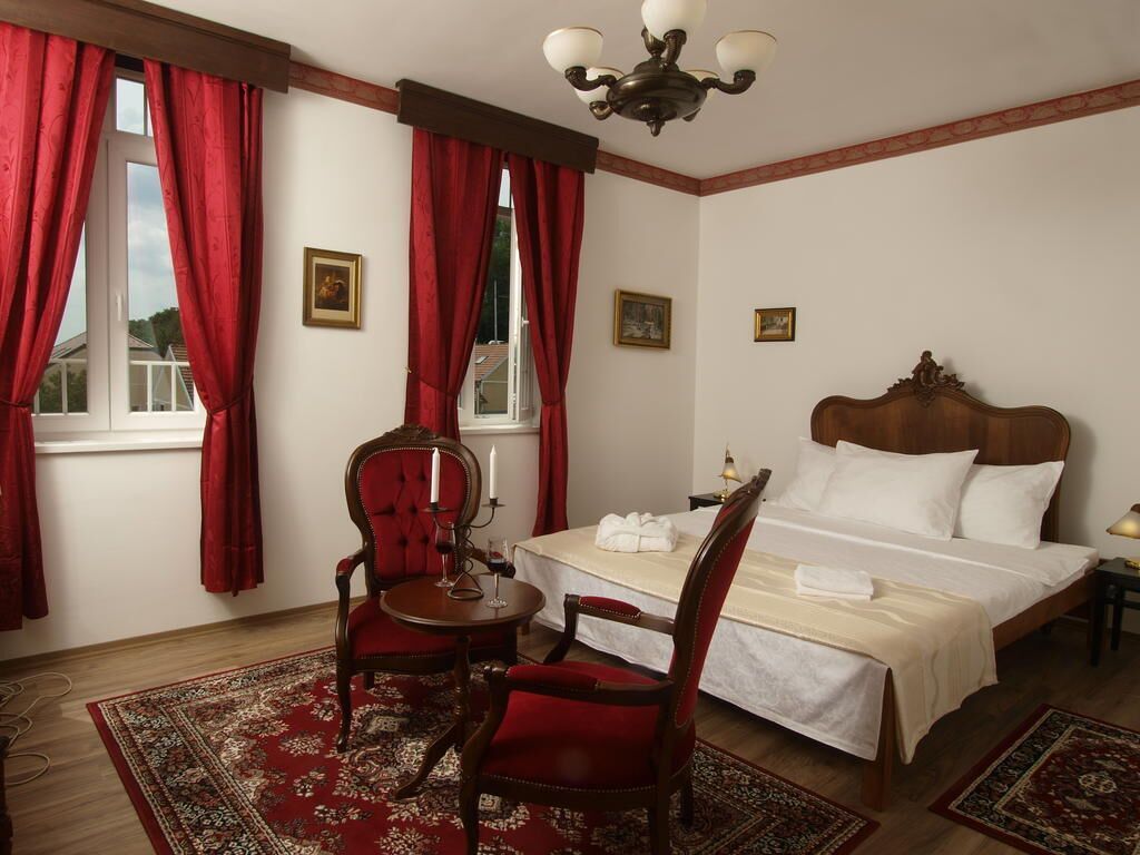 A hotel room with a bed, a chair, a table, and a window with red curtains.