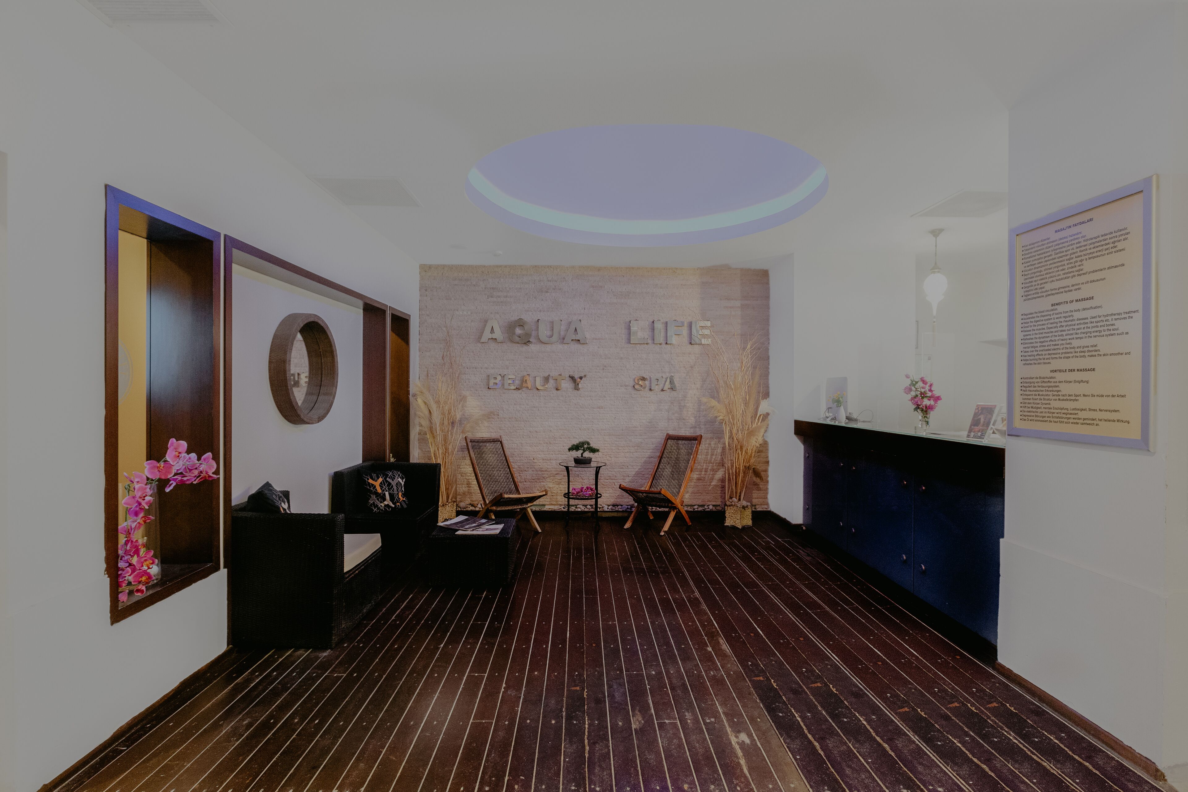 Ein Zimmer mit Holzboden, zwei Stühlen, einem kleinen Tisch mit einer Pflanze und einer Wand mit dem Schriftzug 'AQUA LIFE BEAUTY SPA'.