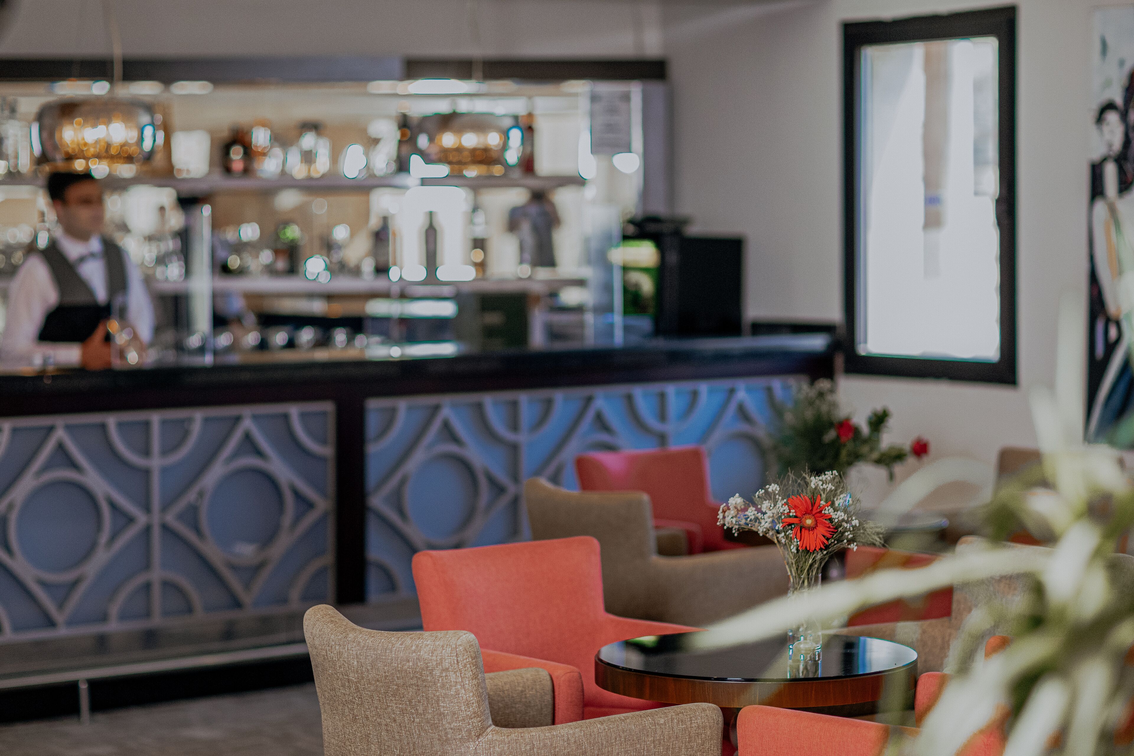 A bar area with a bartender, a counter with various bottles, and seating with a round table and chairs.
