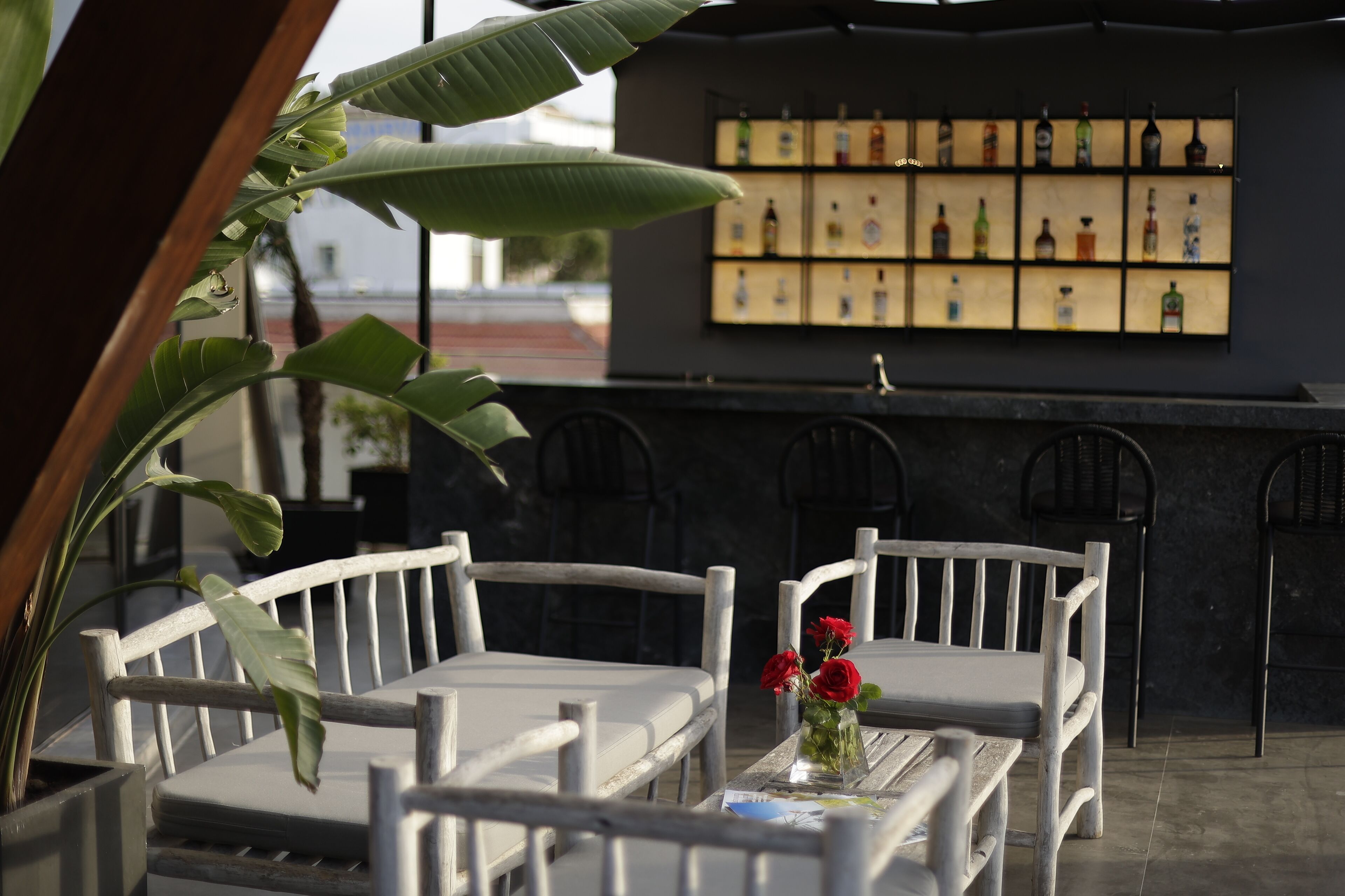 A bar area with a shelf of liquor bottles, white wooden chairs, and a table with a vase of red flowers.