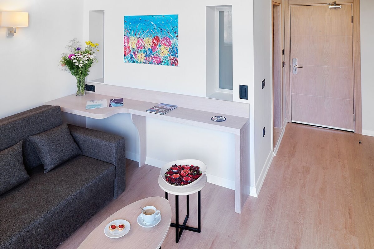 A modern hotel room with a loft bed, a dining area with a table and chairs, a sofa, and a reception desk.
