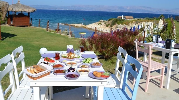 A table set outdoors with a view of the sea, surrounded by greenery and a thatched-roof structure.