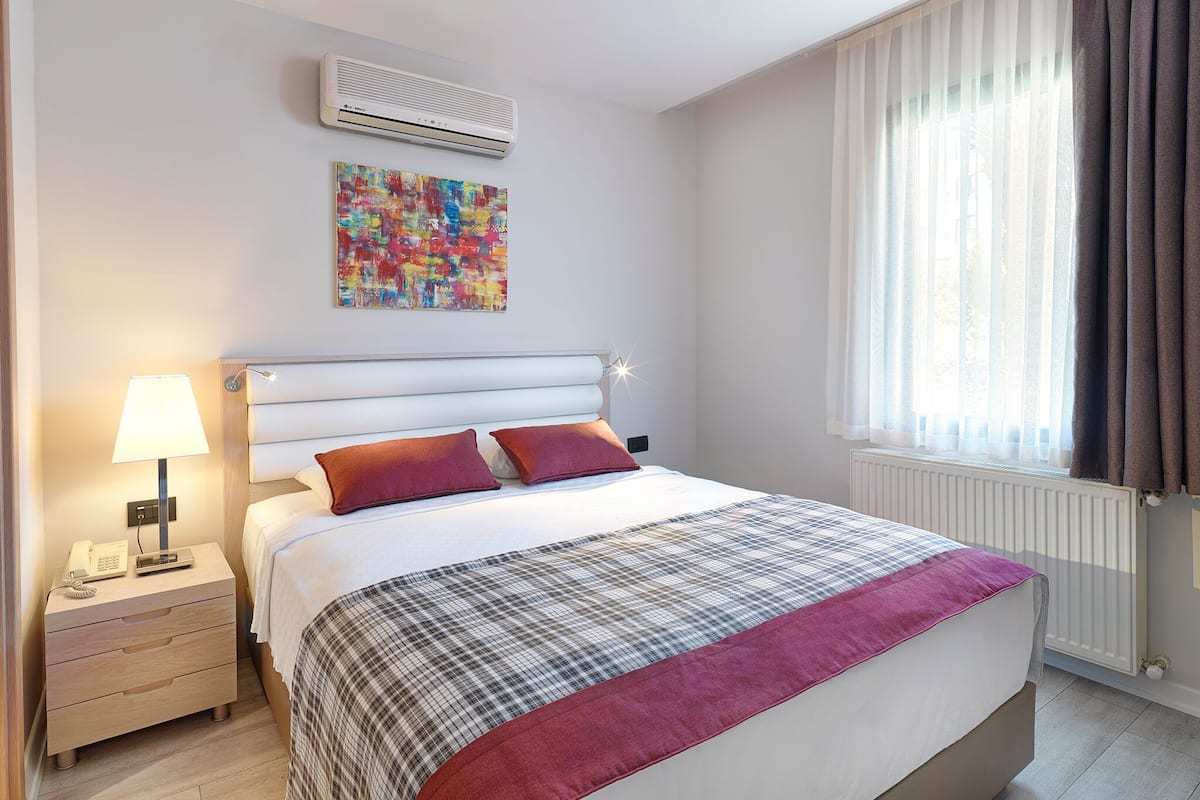 A hotel room with a bed, a bedside table with a lamp, an air conditioner, a colorful abstract painting, and a window with curtains.