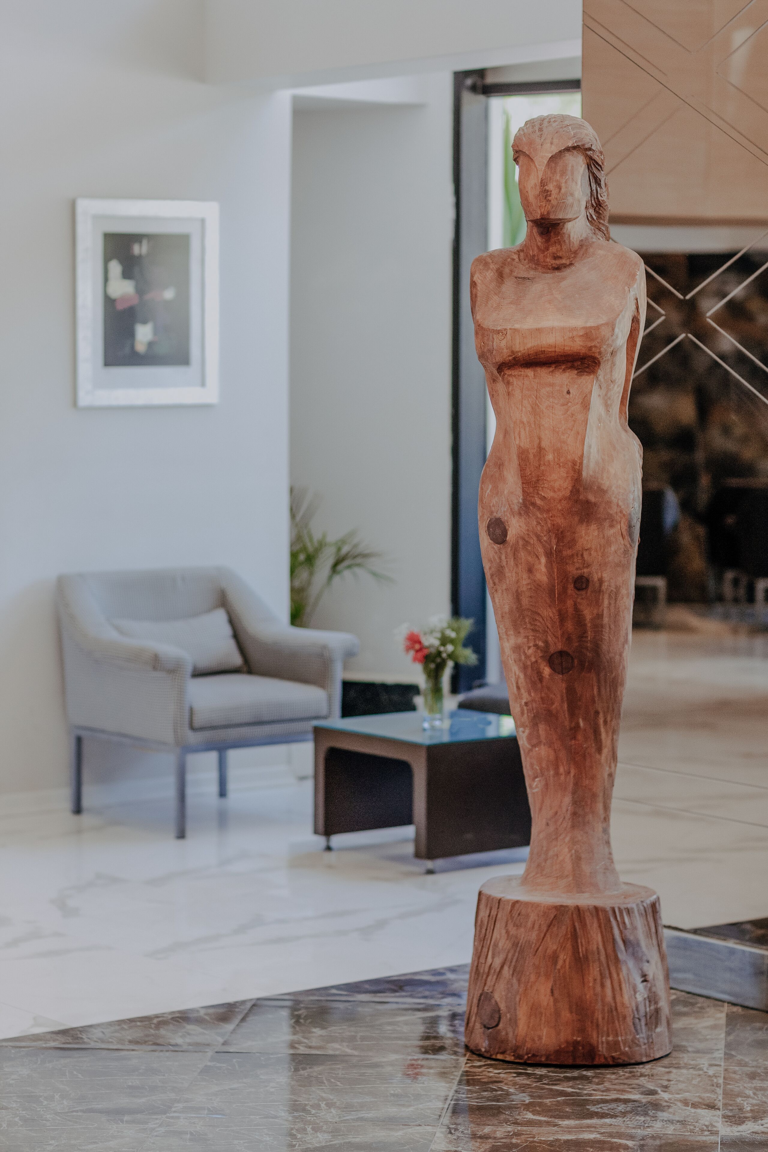Una escultura de madera en un espacio interior moderno, con una silla y una pequeña mesa al fondo.