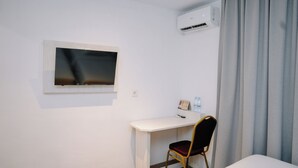 Une chambre d’hôtel avec un bureau blanc, une chaise, un téléviseur à écran plat, un climatiseur, une petite table avec un cadre photo et des bouteilles d’eau, et un rideau gris.