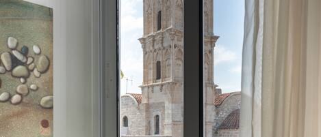 A room with a window overlooking a historic building, a lamp on a bedside table, and a painting on the wall.