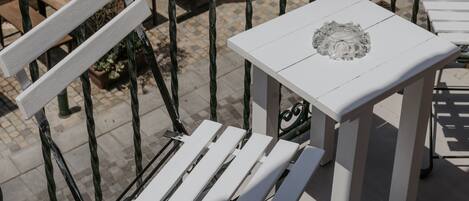 A balcony with white chairs and a table, overlooking a paved area with outdoor seating and umbrellas.