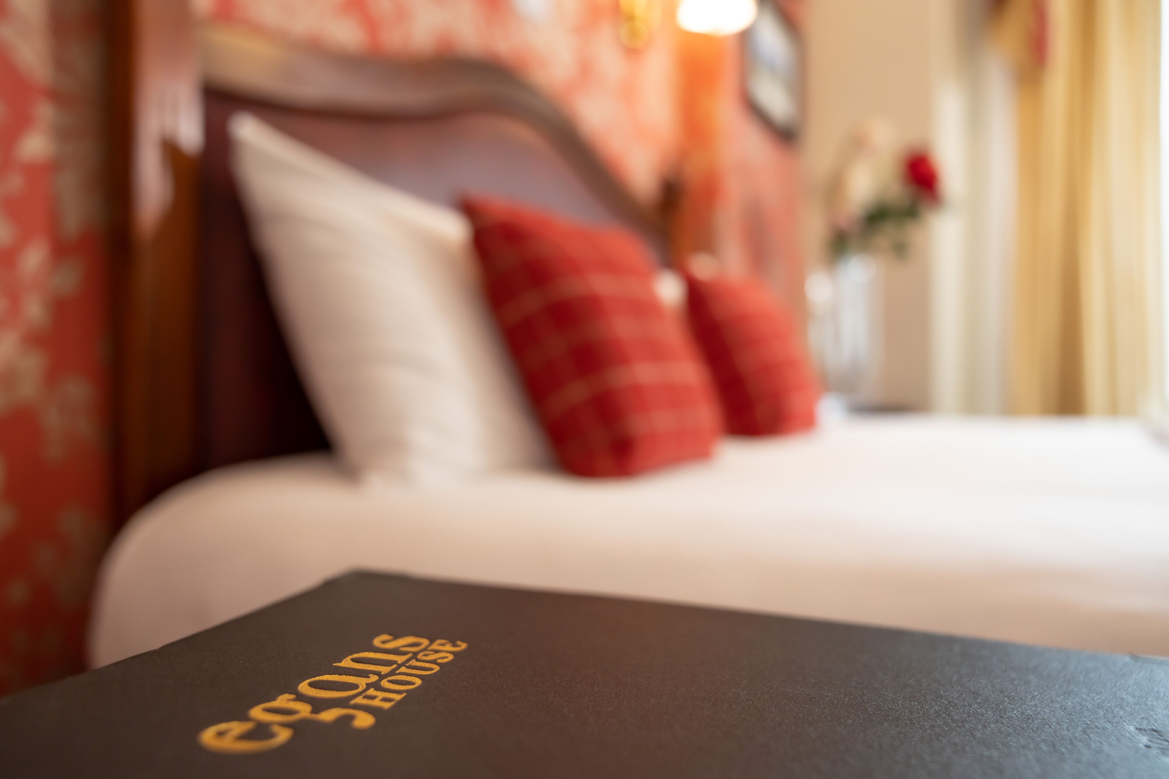 A bed with white linens and red pillows, a dark-colored table with text, and floral wallpaper in the background.