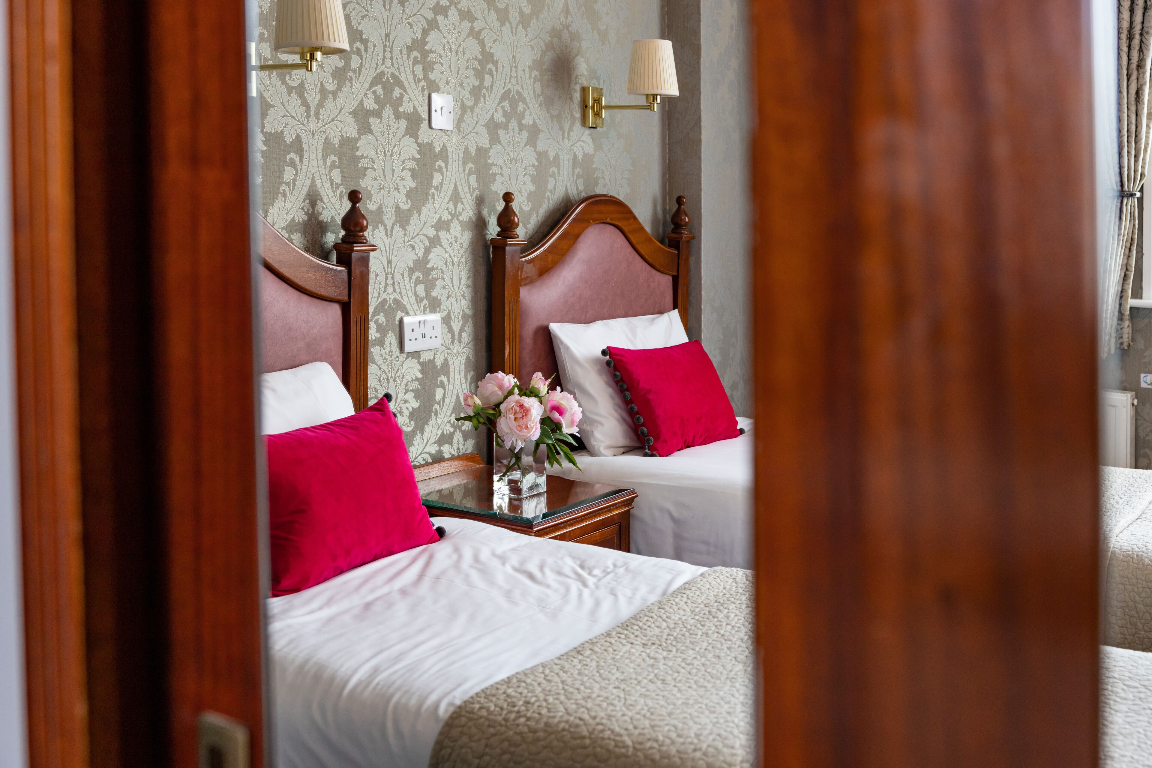 A hotel room with two beds, each with a wooden headboard, white bedding, and red and white pillows. There is a small table with a vase of flowers between the beds.