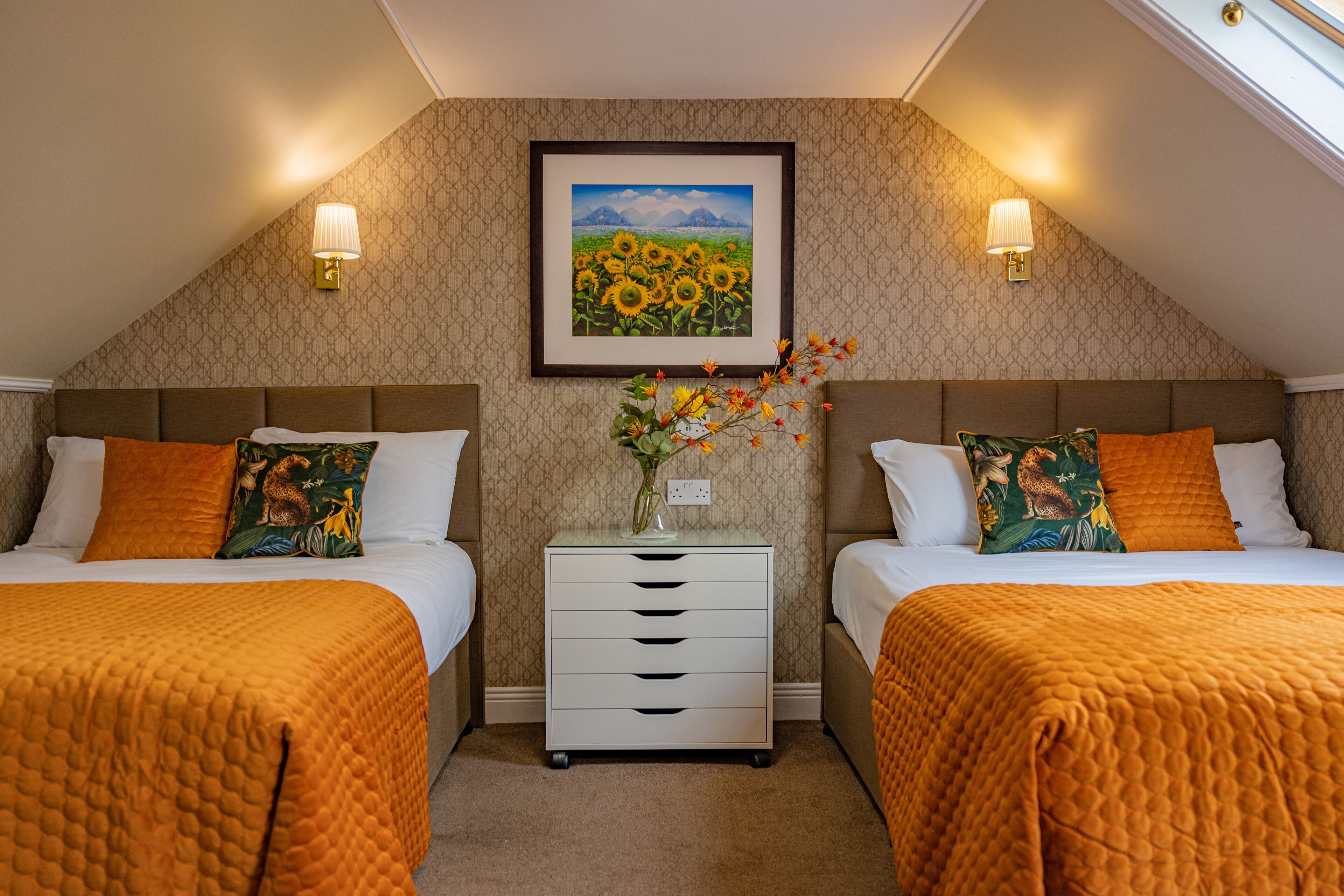 Ein Hotelzimmer mit zwei Betten, einem Nachttisch, einem Bilderrahmen mit Sonnenblumen und Wandleuchten.